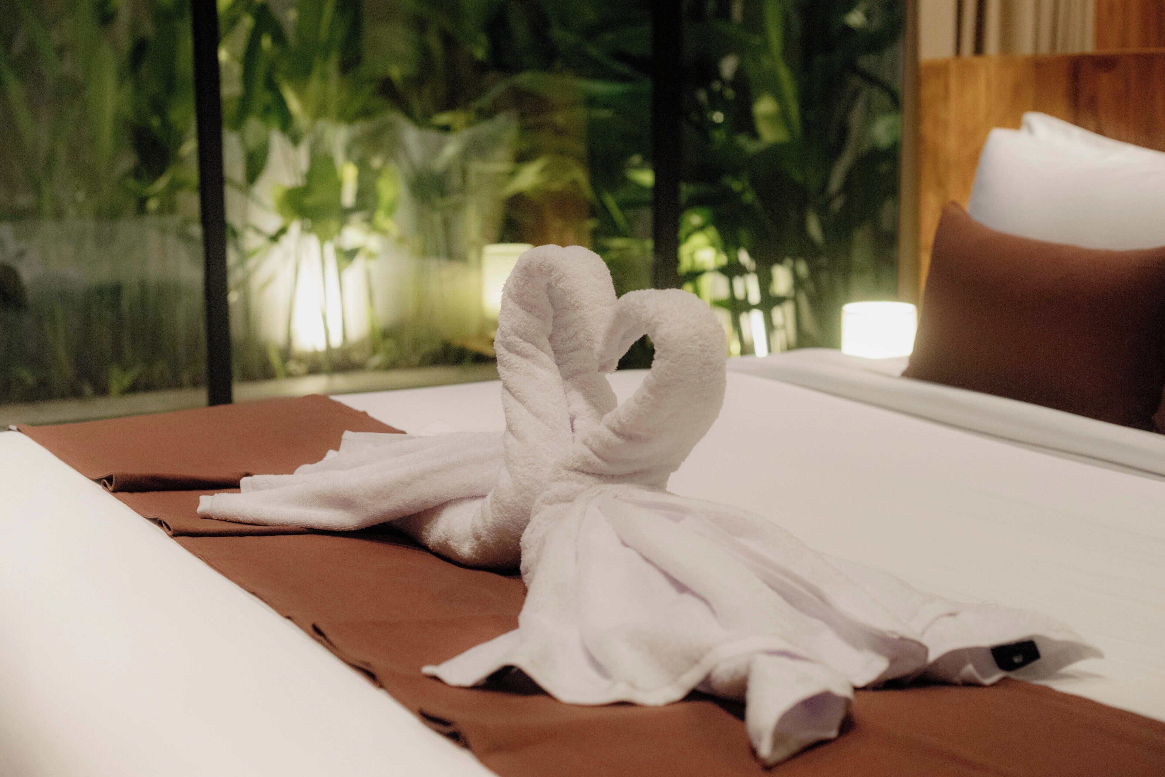 A bed with a towel arranged in a heart shape, a brown blanket, and a white pillow.