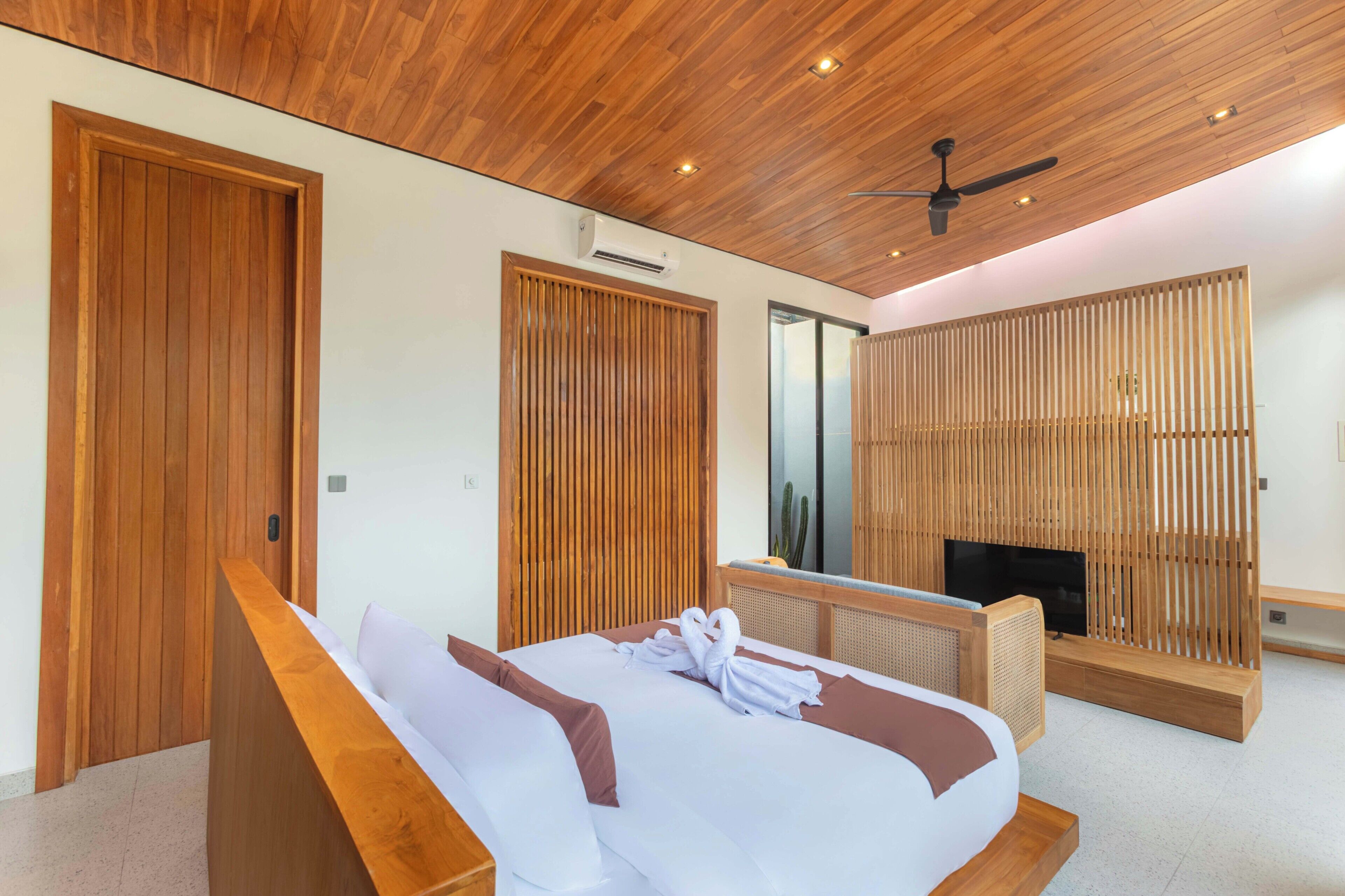A modern hotel room with a wooden ceiling, a bed with white and brown bedding, a TV, and a wooden partition.