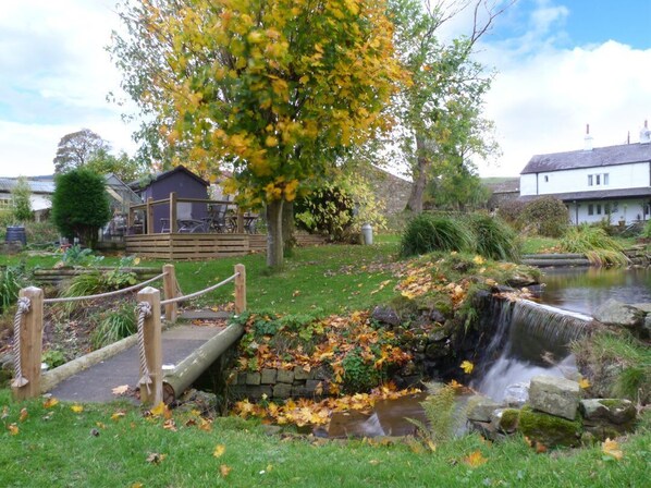 Un jardín con una cascada pequeña, un puente de madera y un edificio al fondo.