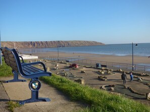 Eine Bank am Meer, ein Sandstrand und ein felsiges Ufer mit Menschen.