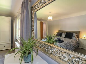 A bedroom with a large mirror, a bed with patterned bedding, a bedside table with a lamp, and a potted plant on a dresser.