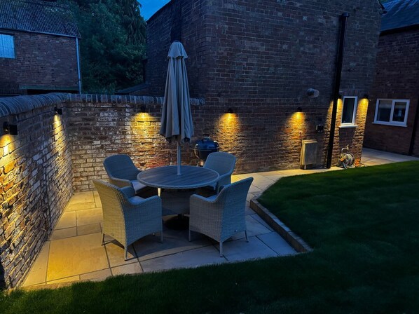 An outdoor patio with a round table and chairs, a barbecue grill, and a brick wall with lights.