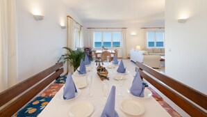 Un espace repas avec une longue table dressée pour le repas, un banc en bois et une vue sur la mer.