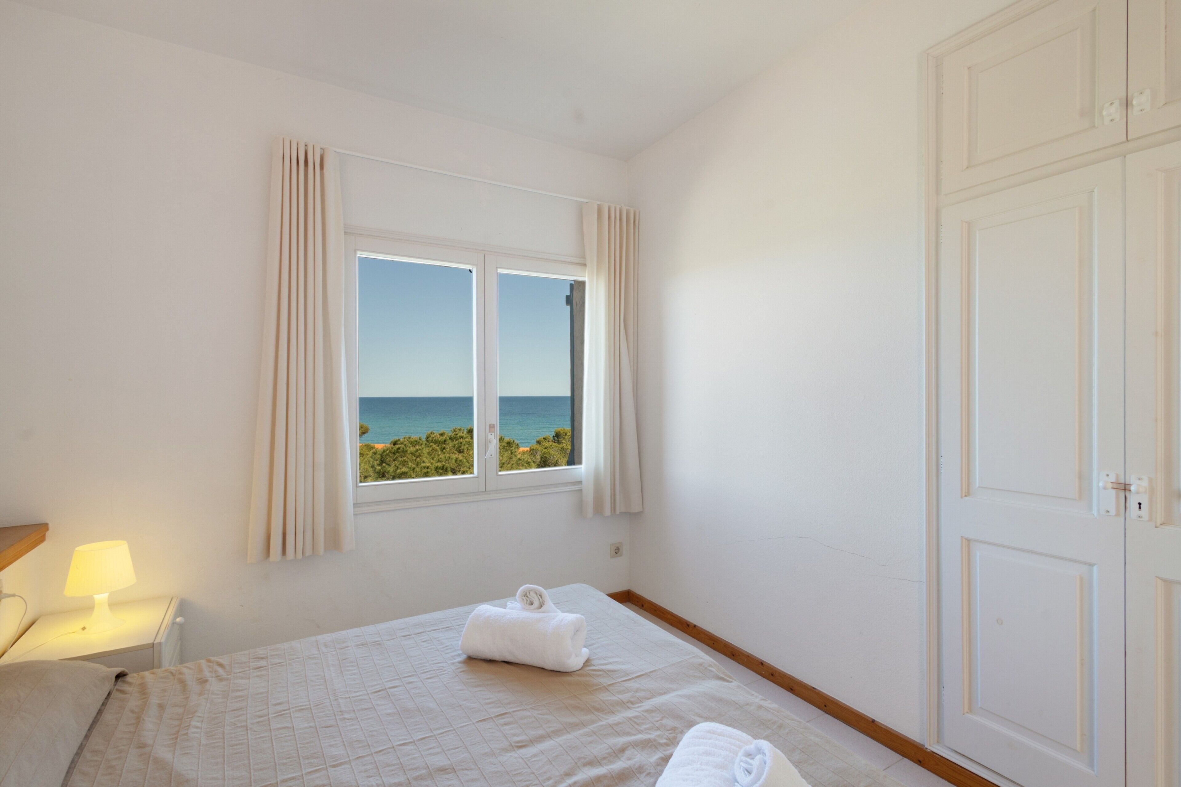 Een slaapkamer met een bed, een nachtkastje, een raam met gordijnen en uitzicht op zee.
