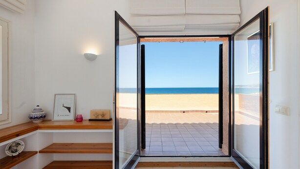 Une pièce dotée d’une porte vitrée donnant sur la plage, d’un plancher en bois, d’une petite étagère ornée d’objets décoratifs et d’un mur blanc.