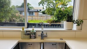 A kitchen with a double sink, a window with a view of a backyard, and a potted plant on the windowsill. - Sunny 3BR Home - Close to Essentials & Transport (Auckland)
