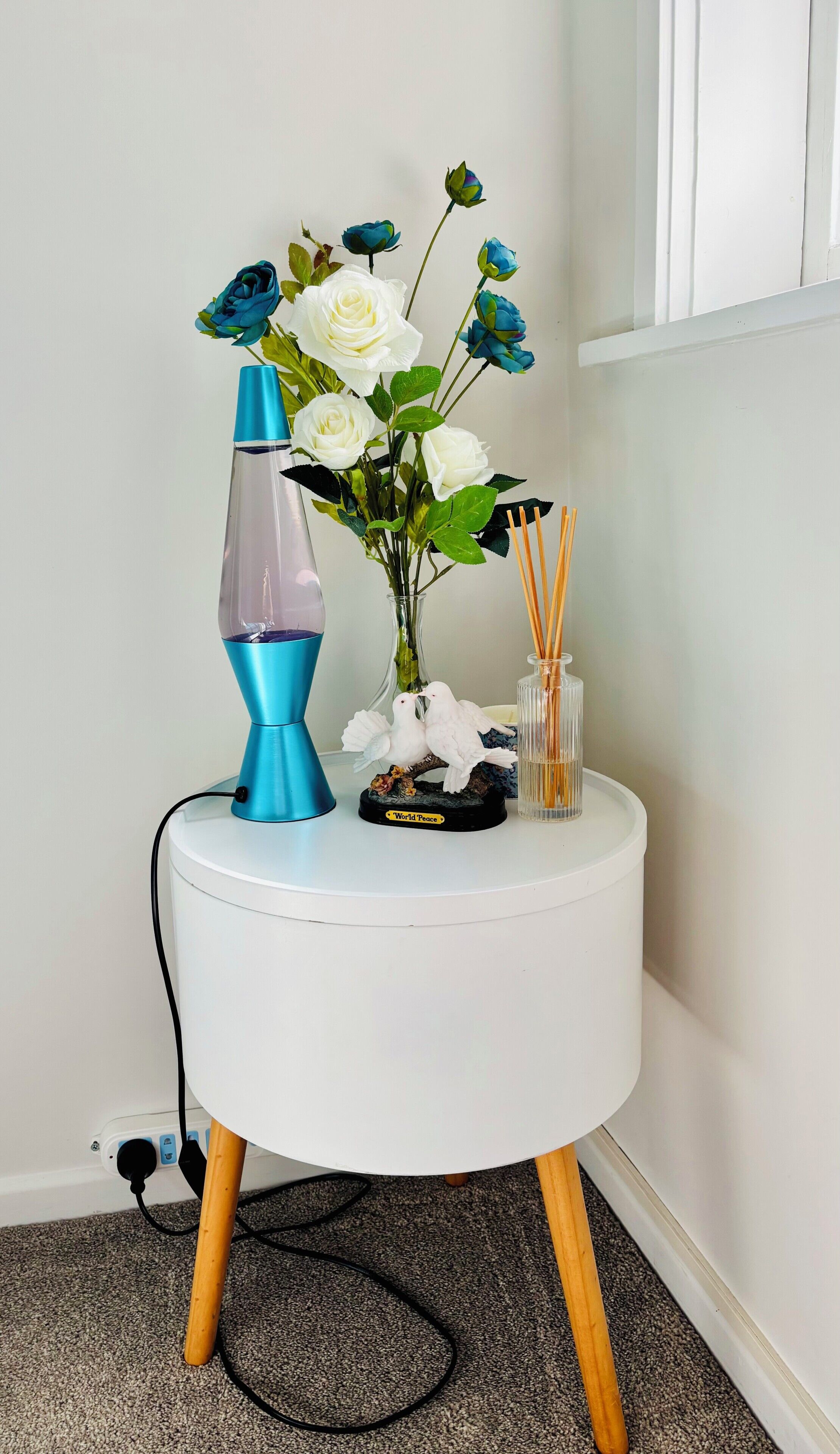 A white round side table with a blue lava lamp, a vase with white and blue flowers, a 'World Peace' bird figurine, and a container with reed diffusers.