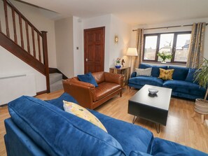 A living room with a brown leather sofa, blue sofas, a coffee table, a wooden staircase, and a large window with curtains. - Sylvannah, Buxton, Derbyshire. (Buxton)