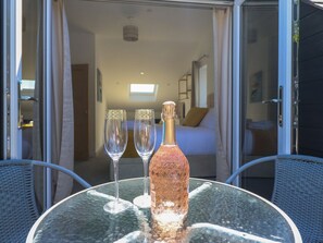 Ein Glastisch mit einer Flasche und zwei Gläsern, ein Korbstuhl und ein Blick in einen Raum mit einem Bett und einem Fenster.