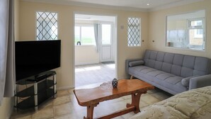 A living room with a TV, sofa, and table, a balcony with a view, and a door leading outside. - Watersmeet - 85 @ Park Avenue Holiday Village (Sheerness)