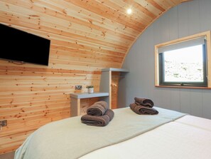 Ein Zimmer mit Holzboden, einem Bett mit weißer Matratze, zwei gerollten Handtüchern, einem an der Wand montierten Flachbildfernseher und einem Fenster mit Blick auf Grünflächen.