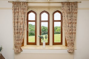 Ein Zimmer mit einem großen Fenster, das drei Rundbogenfenster und bunte Vorhänge sowie einen weißen Vasen mit Grünpflanzen auf dem Fensterbrett zeigt.