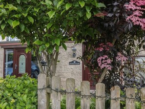 Ein Holzzaun mit Stacheldraht, ein Steinhaus mit einem Schild, auf dem 'FOXGLOVES' steht, und ein Baum mit rosafarbenen Blüten.