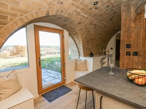 A cozy room with a curved ceiling, a wooden bench, a glass door leading outside, and a kitchen area with a bar stool and fruit bowl. - Tan y Wawr- Onnen, Llanfair Caereinion (Welshpool)