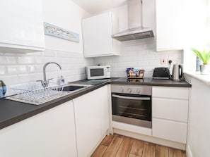 A compact kitchen with white cabinets, a microwave, an oven, and a black countertop. - Sandy Shell Cottage, Conwy (Conwy)