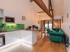 A modern kitchen with white cabinetry, a built-in microwave, and a green backsplash. There is a dining area with a green sofa and a wooden table. The room has wooden beams on the ceiling and a large window. - Repton @ Engine Shed - Whitby Retreat (Whitby)