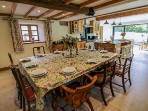 Ein Essbereich mit einem langen Tisch, gedeckt für sechs Personen, Holzsesseln, einer Tischdecke mit Blumenmuster und Blick auf die Küche und den Außenbereich.