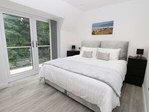 Ein Schlafzimmer mit einem großen Bett, einem Nachttisch, einem Tischchen, einem Bild an der Wand und einer Glastür, die auf einen Balkon führt.