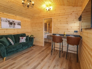 A wooden cabin interior with a green sofa, two bar stools, a dining table, a chandelier, and a painting of horses on the wall. - The Stable, Church Broughton (Derby)