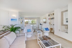 A spacious living area with a sofa, wicker chairs, a glass coffee table, and a view through a large window.