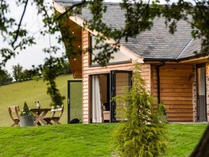 Une cabane en bois avec un toit en pente, un coin repas avec des sièges à l’extérieur et un petit arbre au premier plan.