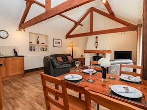 Een eethoek met een houten tafel voor vier personen, een zwarte leren bank, een open haard en een keuken met houten kastjes en een klok.