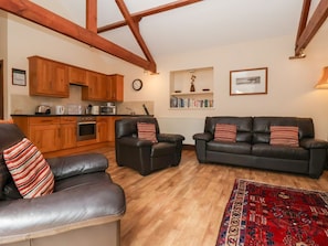 Een woonkamer met een bank, twee fauteuils, een salontafel, een keuken met houten keukenkastjes, een klok en een boekenplank.