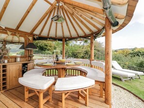 Ein hölzerner Pavillon mit einem runden Tisch und weißen, gepolsterten Stühlen, umgeben von einer Kiesfläche und einem Rasen.