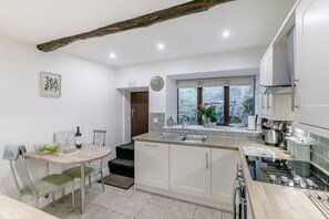 A modern kitchen with white cabinetry, a dining area with a round table and chairs, a window with a view, and a rustic ceiling beam. - Weavers Cottage, Heptonstall (Hebden Bridge)