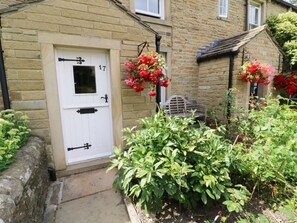 A stone house with a white door marked '17', a small garden with green shrubs, and hanging flower baskets. - Sally's Cottage, Carleton-In-Craven (Skipton)