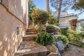 A stone staircase leading to a white building with a balcony, surrounded by greenery and palm trees.