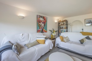 A modern hotel room with two white sofas, a round coffee table, a bookshelf, and a large painting on the wall.