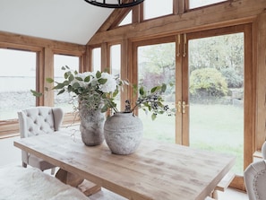 Une table à manger en bois, agrémentée de deux grands vases remplis de plantes, d’un fauteuil capitonné et de grandes fenêtres offrant une vue sur la verdure.