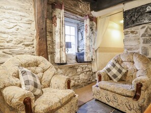 Een rustieke kamer met stenen muren, twee gestreepte fauteuils, een raam met gordijnen en een stenen openhaard.