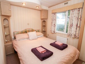 A bedroom with a bed, two pillows, a window with curtains, a radiator, and a shelf with decorative items. - 7 Old Orchard, Much Wenlock (Much Wenlock)