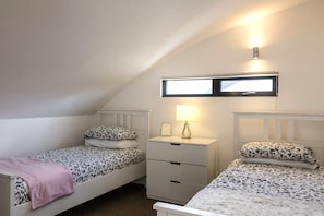 Un alojamiento pequeño y de una sola habitación con dos camas individuales, una mesita de noche con lámpara, una ventana y un aplique de pared.