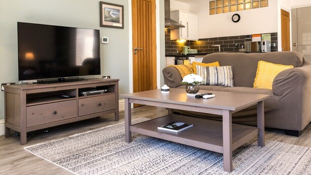 Ein Wohnzimmer mit Flachbildfernseher, Couchtisch, einer Couch mit Kissen sowie einer kleinen Seitentisch und einem Küchenbereich im Hintergrund.