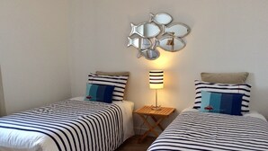 A room with two beds, striped bedding, a wall-mounted mirror, a small table with a lamp, and a wooden floor. - Baie des Veys Cottage near Utah Beach (Brucheville)