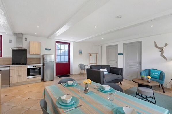 A modern kitchen with a stove, oven, and refrigerator, a dining area with a table set for two, and a living room with a sofa and a decorative wall piece.
