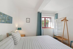 A bedroom with a bed, a bedside table, a lamp, a window with curtains, a radiator, and a wooden drying rack.