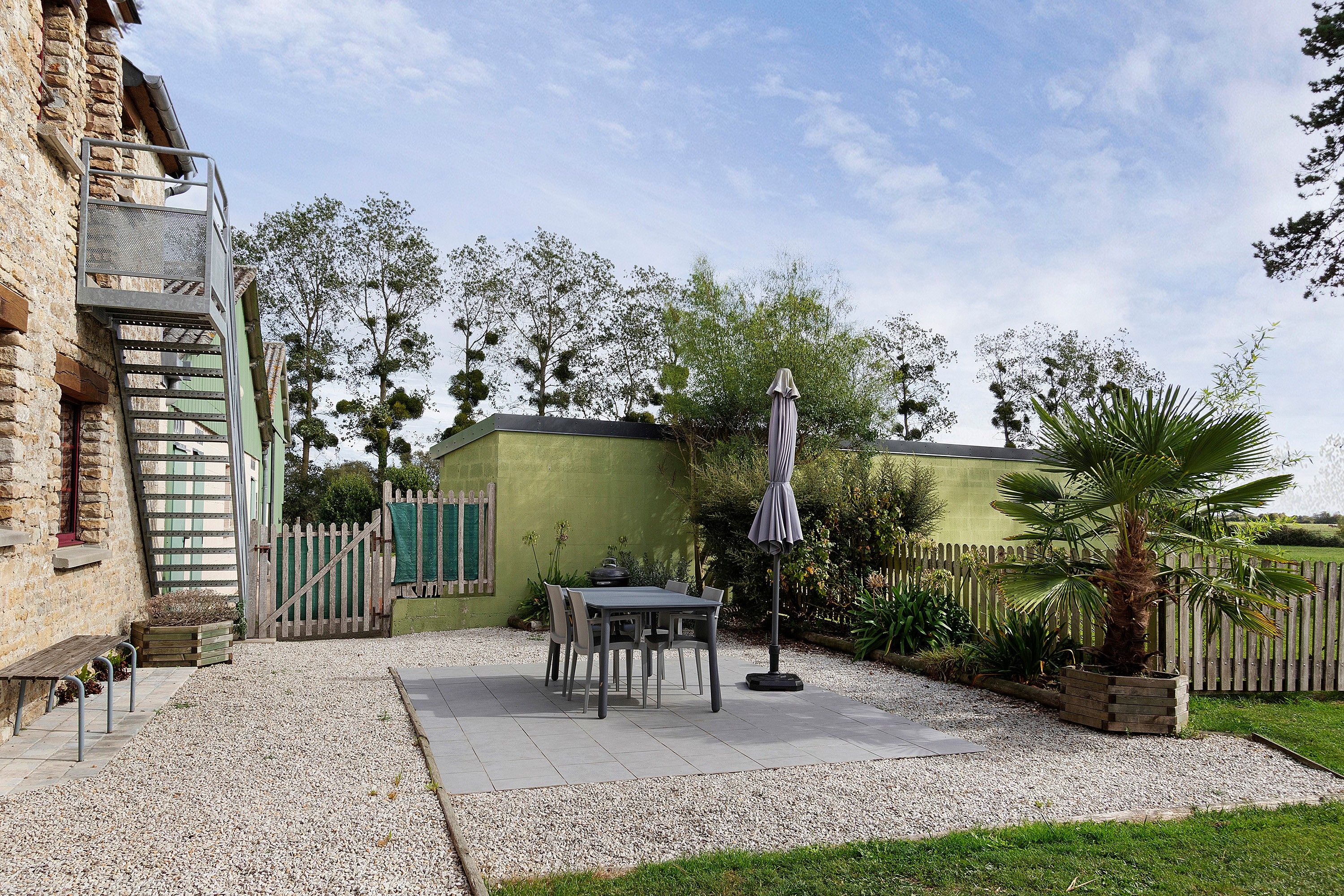 Eine Terrassenfläche mit Tisch und Stühlen, ein kleiner Garten mit einer Palme und ein Gebäude mit grüner Wand und Holzzaun.