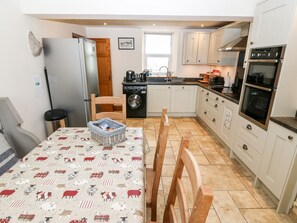 A compact kitchen with a washing machine, a table with a patterned tablecloth, and wooden chairs. - Cefn Werthyd Farmhouse Retreat (Caernarfon)