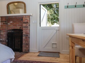 Ein Zimmer mit Kamin aus Backsteinen, einer weißen Tür mit diagonalem Muster, einem kleinen Fenster und einem Holztisch.