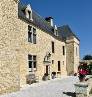 Un edificio in pietra con uno stile architettonico tradizionale, caratterizzato da un ingresso centrale con un arco decorativo, numerose finestre e un vialetto sterrato.