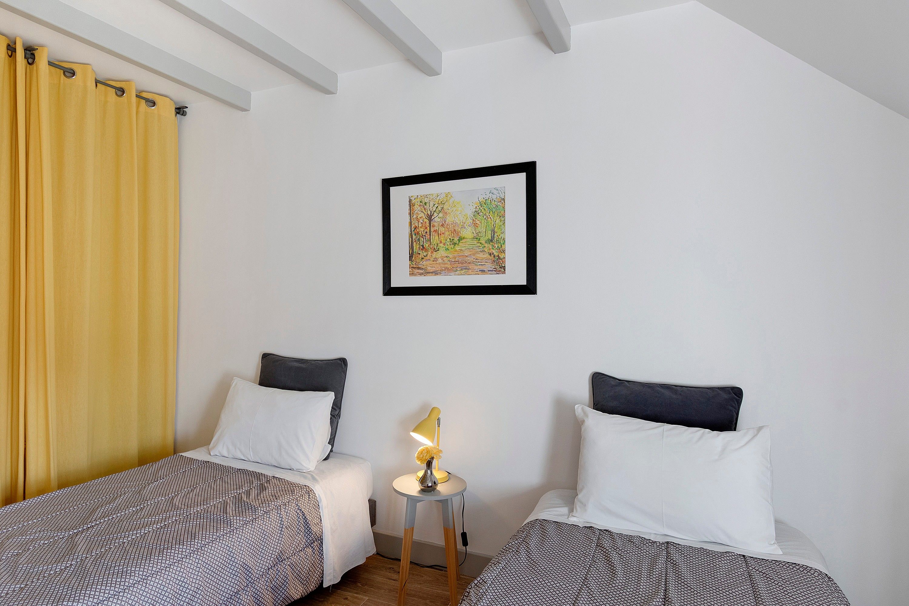 A room with two beds, a small table with a lamp, and a framed picture on the wall.