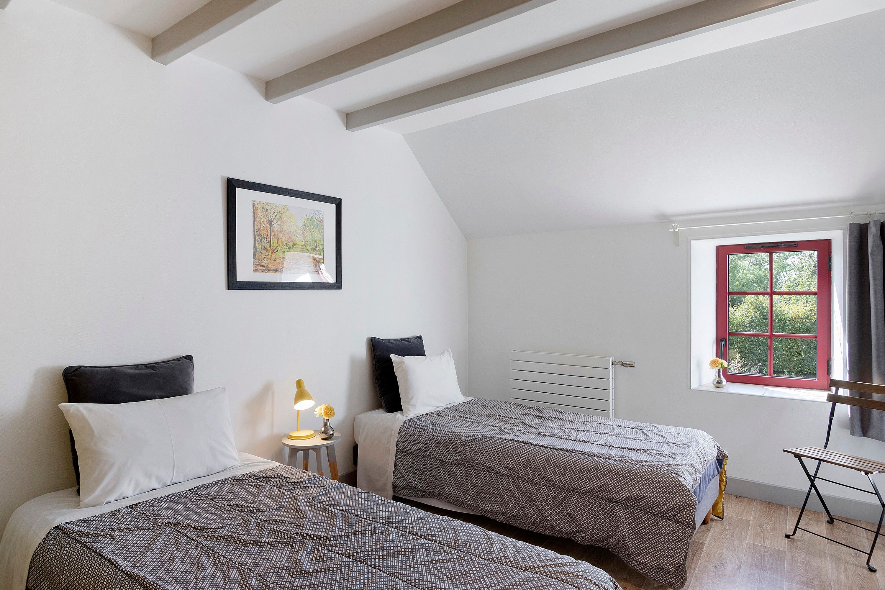 Ein Schlafzimmer mit zwei Betten, einem Fenster mit rotem Rahmen, einem Heizkörper und einem gerahmten Bild an der Wand.