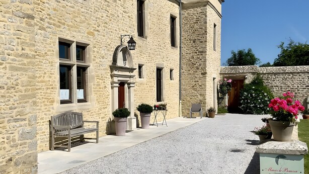 Un bâtiment en pierre avec une allée gravillonnée, un banc, des plantes en pot et un panneau.