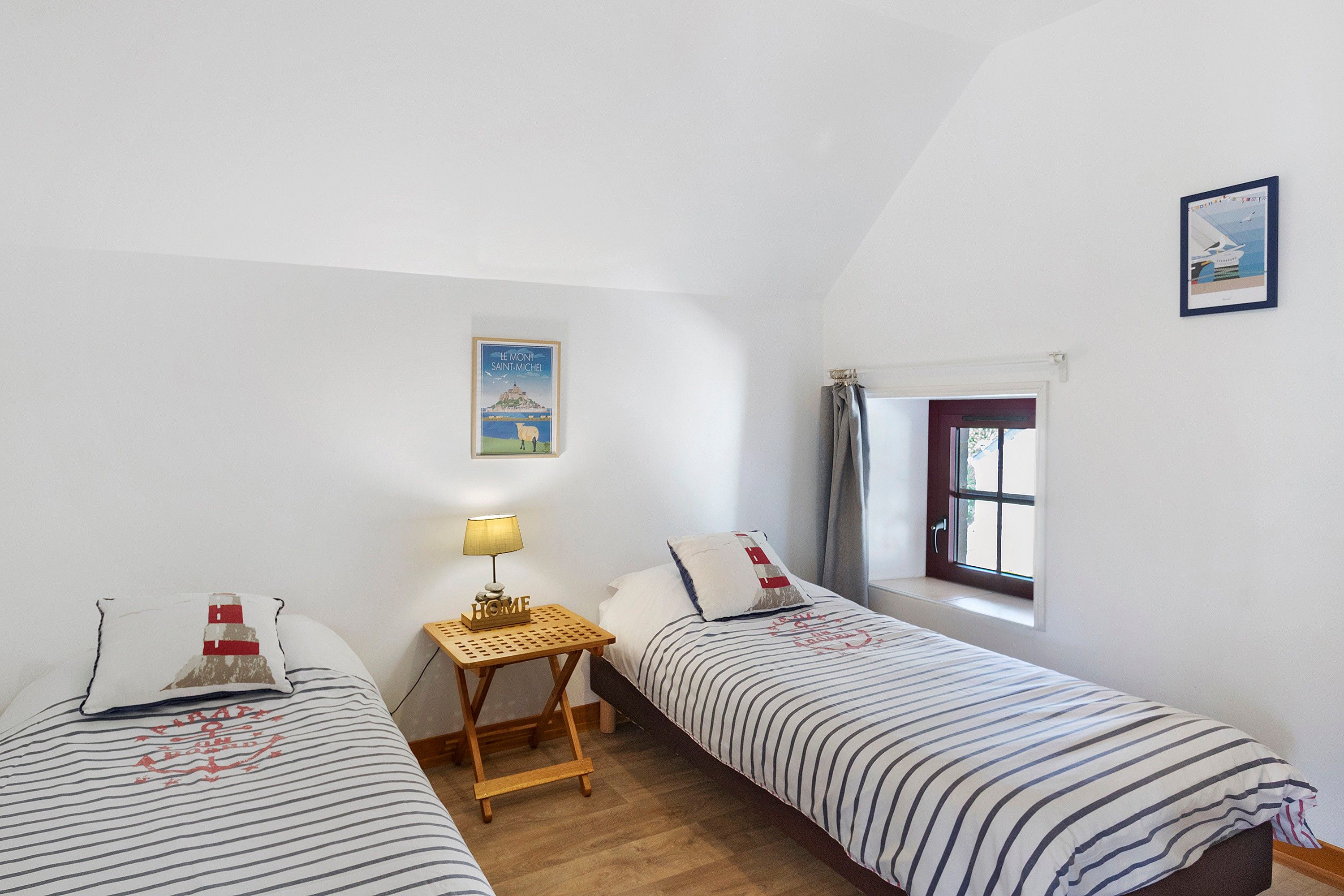 A bedroom with two single beds, a small table with a lamp, a window with curtains, and a wall-mounted picture.