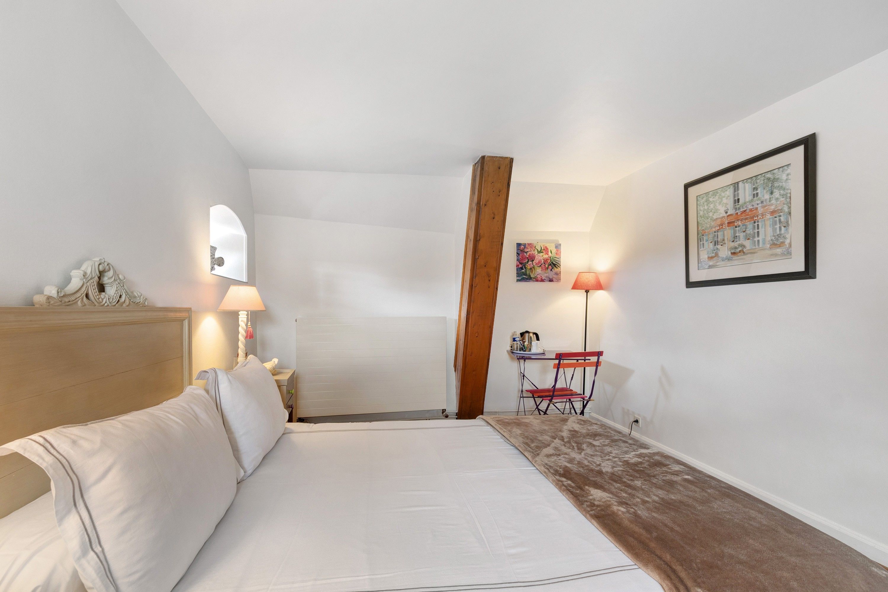 Een slaapkamer met een bed, nachtkastje, lamp, houten balk, stoel, tafel en een schilderij aan de muur.