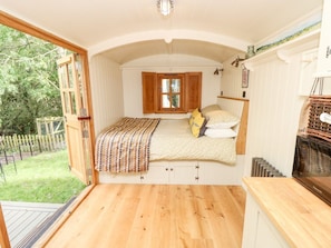 Une chambre avec un plancher en bois, un lit recouvert d’une literie à motifs, une fenêtre avec des volets en bois et une vue sur un jardin.